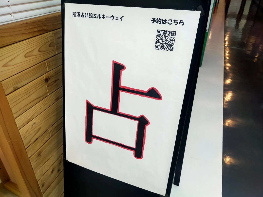 「所R占い館ミルキーウェイ」の看板で、大きく「占」の文字と「予約はこちら」の案内、そして予約用のQRコードが描かれています。