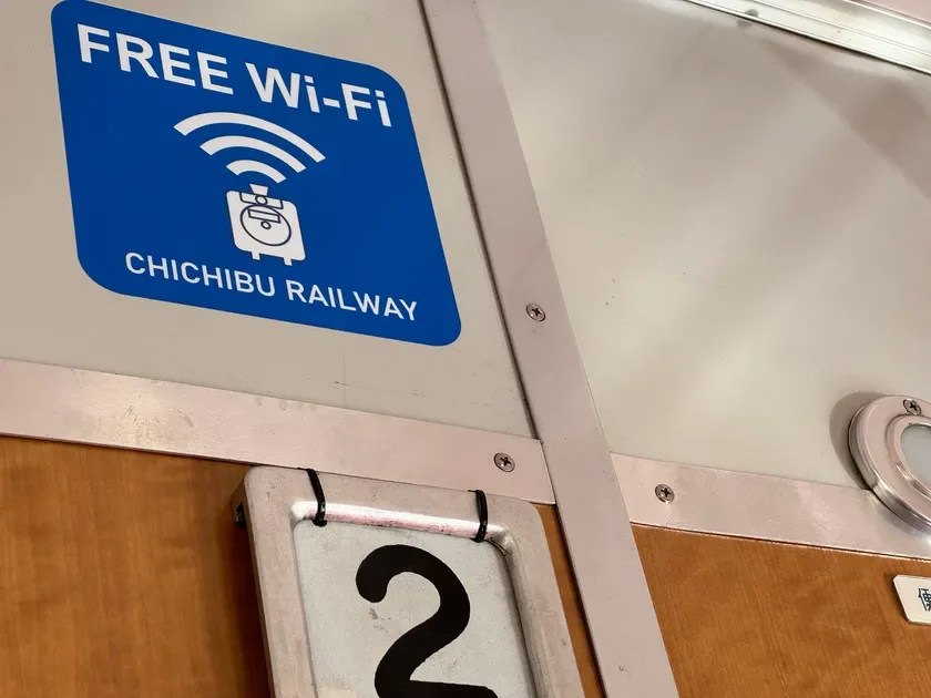 秩父鉄道の車内らしき場所に設置された、無料Wi-Fiサービスを示す青い案内表示です。電波のアイコンとスーツケースのイラストが描かれ、「FREE Wi-Fi」と「CHICHIBU RAILWAY」の文字が読み取れます。一部に「2」の数字も見えます。