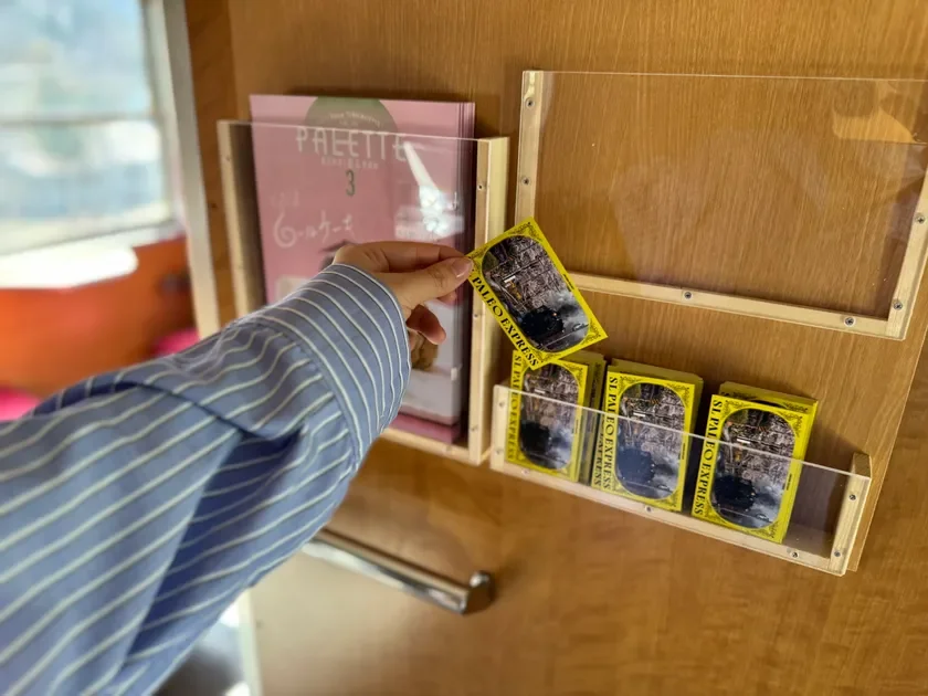 ストライプシャツを着た人物が、列車の車内で「SLパレオエクスプレス」のグッズを手に取っている様子。隣にはパンフレットも置かれており、旅の思い出やお土産を選んでいる瞬間を捉えている。