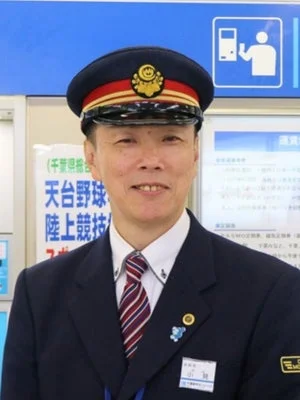 駅の制服を着た中年の男性が笑顔で立っているポートレートです。背景には駅の案内板や標識が見えます。