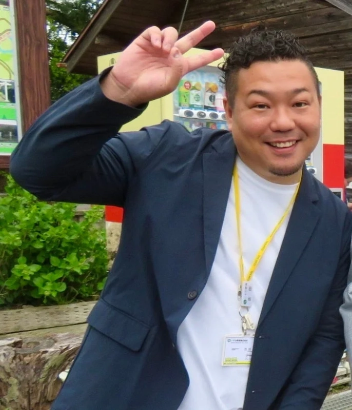 笑顔でピースサインをする男性が写っています。紺色のジャケットと白いTシャツを着用し、首からネームプレートを下げています。背景には緑や自動販売機が見え、屋外でのリラックスした雰囲気が伝わります。