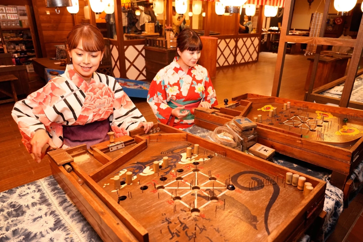 浴衣を着た二人の女性が、レトロな雰囲気の屋内で伝統的なスマートボールのようなゲームを楽しんでいます