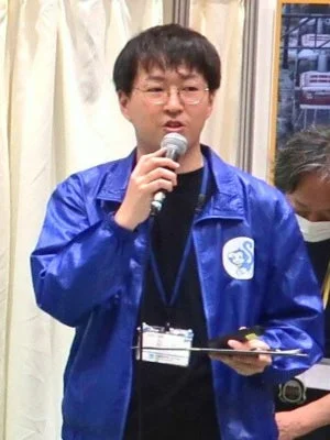 青いジャケットを着た男性がマイクを持って話している様子。眼鏡をかけ、首から名札を下げていることから、イベントでのプレゼンテーションか講演をしている場面と推測されます。ジャケットには猿のロゴが見えます。