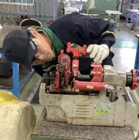 工場のような環境で、作業員が配管用のネジ切り機を操作している様子。作業員は身を乗り出し、機械の赤いハンドル部分を注意深く確認しながら作業を進めている。