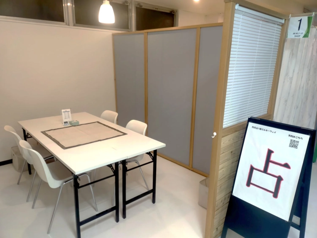 白いテーブルと椅子が並び、パーテーションで区切られた室内空間です。「所沢占い館ミルキーウェイ」の看板があり、「占」の文字が目立ちます。