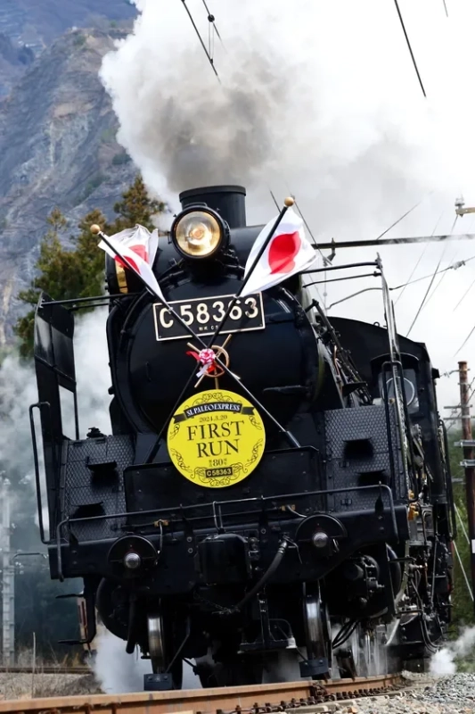 日本の蒸気機関車C58363「SLパレオエクスプレス」が、2021年3月20日のファーストランで力強く煙を上げながら走行する様子。日の丸が飾られ、山を背景にした特別な運行を捉えた一枚です。
