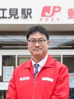 赤いジャケットと眼鏡をかけた中年の日本人男性が、江見駅と日本郵便のロゴが見える建物の前で立っています。郵便局員と思われる男性がカメラに向かって笑顔を見せている様子です。