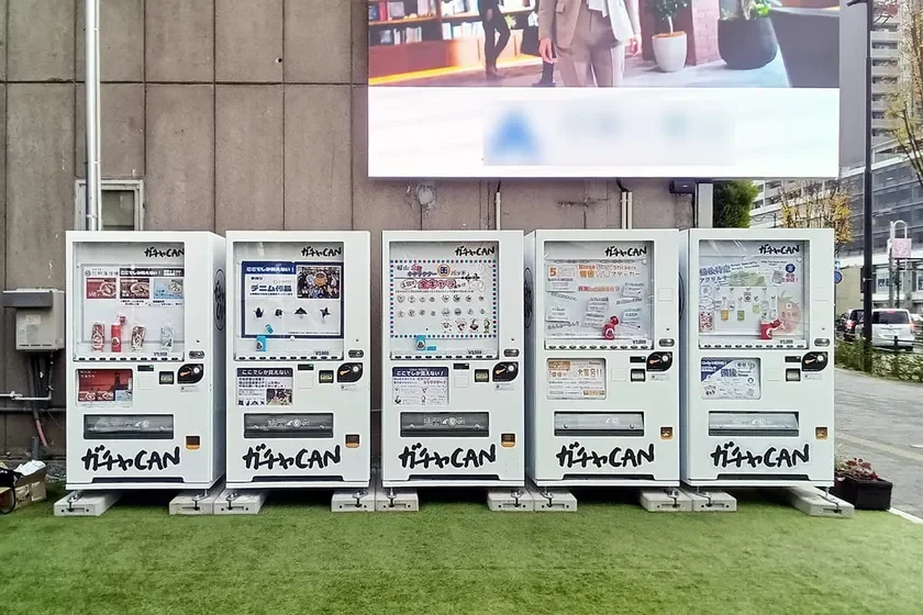 人工芝の上に「ガチャ・CAN」と書かれた白い自動販売機が5台並んでいます。