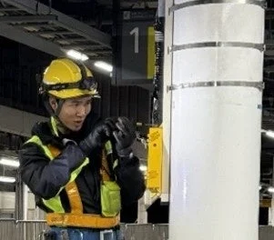 黄色いヘルメットと安全ベストを着用した作業員が、屋内の駅のような場所で手元の作業に集中している様子。背景には「1」の標識が見えます。