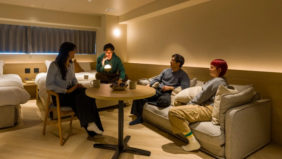 温かい照明の部屋で、男女4人がソファや椅子に座り、テーブルを囲んで談笑している様子