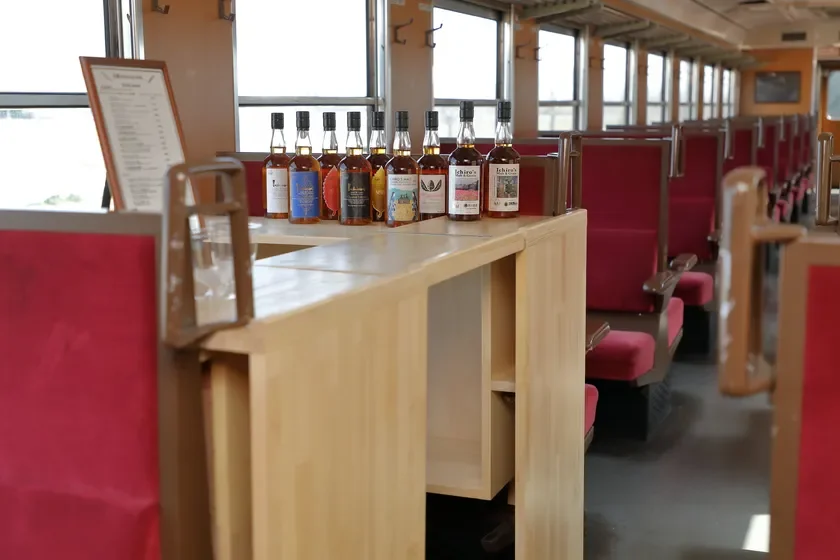 列車内で木製のカウンターに「Ichiro's Malt」のウイスキーボトルが複数並べられています。赤い座席が続く車内の様子から、鉄道でのイベントや特別車両での提供風景を捉えた一枚です。