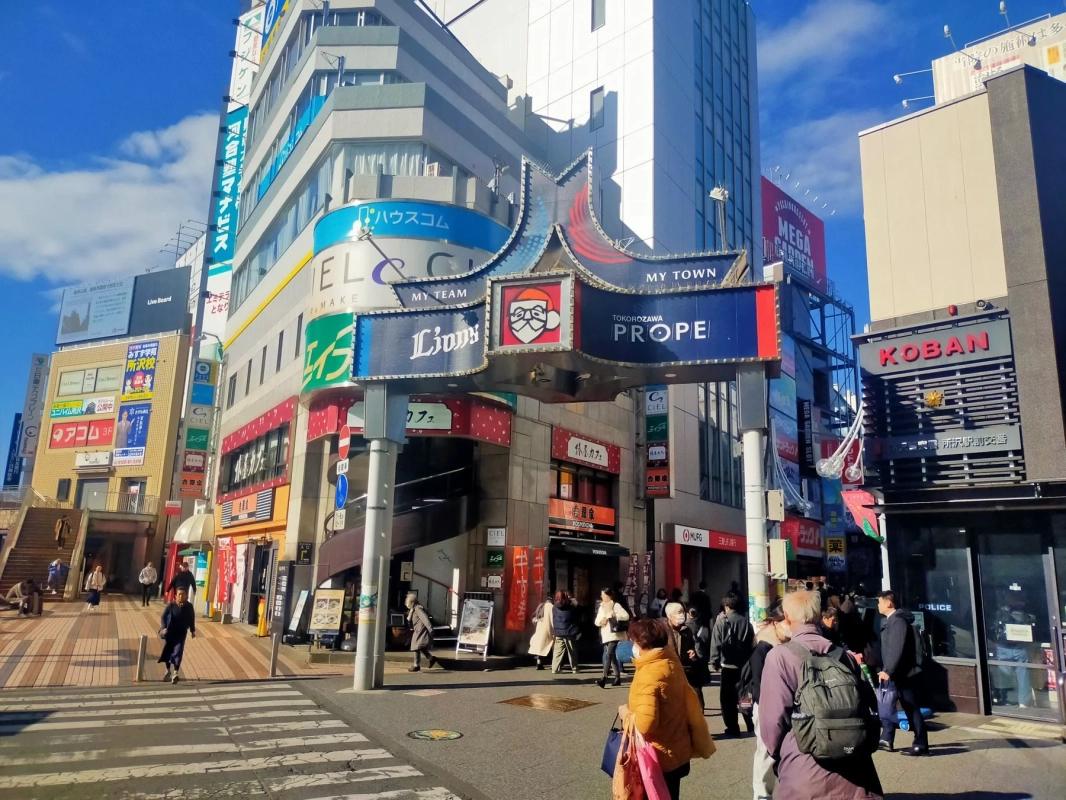 晴れた日の日本の賑やかな駅前商店街の風景。多くの人々が行き交い、様々な店舗の看板が並ぶ商業ビルが立ち並ぶ。