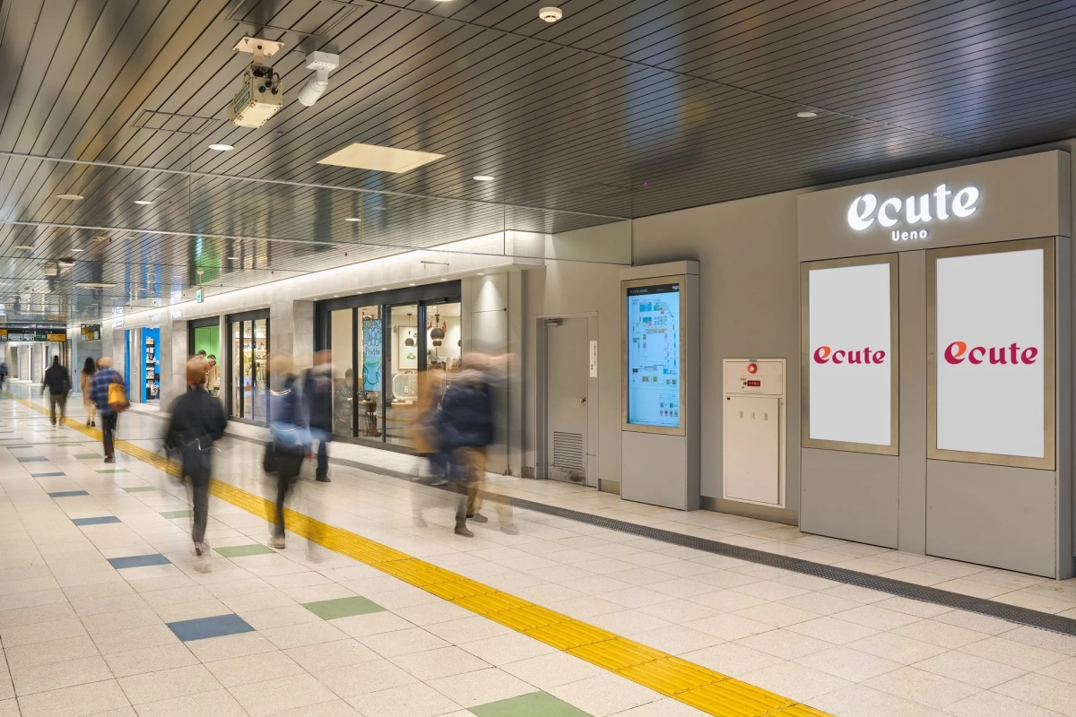 駅構内と思われる地下通路で、多くの人々が歩いています。ecute上野の看板や店舗、デジタル式のフロアガイドが見られ、床には点字ブロックが設置されています。