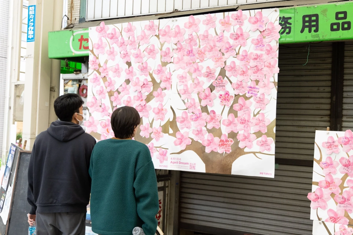 街角に設置された、桜の花びら型のメッセージが多数貼られた大きな掲示板を、マスクをつけた人々が見上げている様子。花びらには「NO WAR」や「世界平和」といった願い事や夢が書かれており、「April Dream」という企画の一環と見られる温かい光景です。