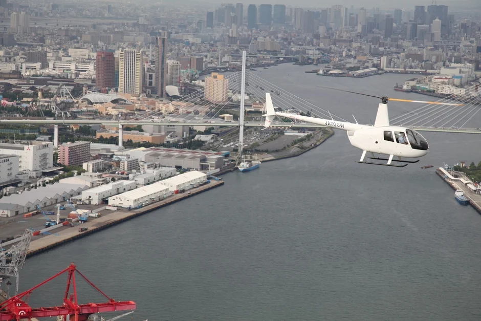 白いヘリコプターが、高層ビル群や吊り橋が広がる湾岸都市の上空を飛行している様子です。