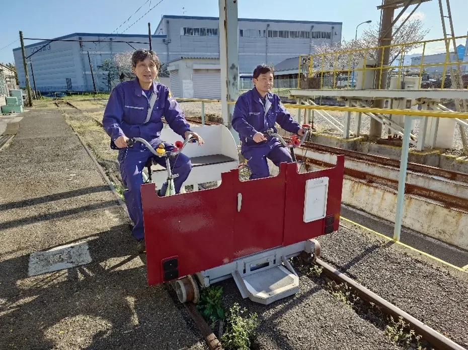 青い作業着を着た2人の男性が、鉄道の線路の上をレールバイクに乗って笑顔で進んでいます
