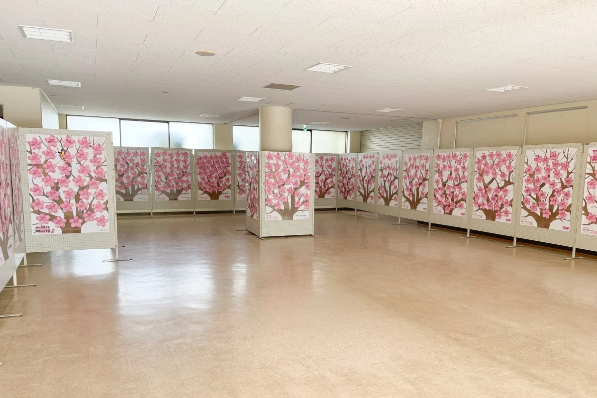 広い室内に桜の木が描かれたパネルが多数展示されています。桜の花びらの部分には来場者からのメッセージが貼られており、希望や願いが込められたイベントの様子がうかがえます。特に「April Dream」という文字が見えます。