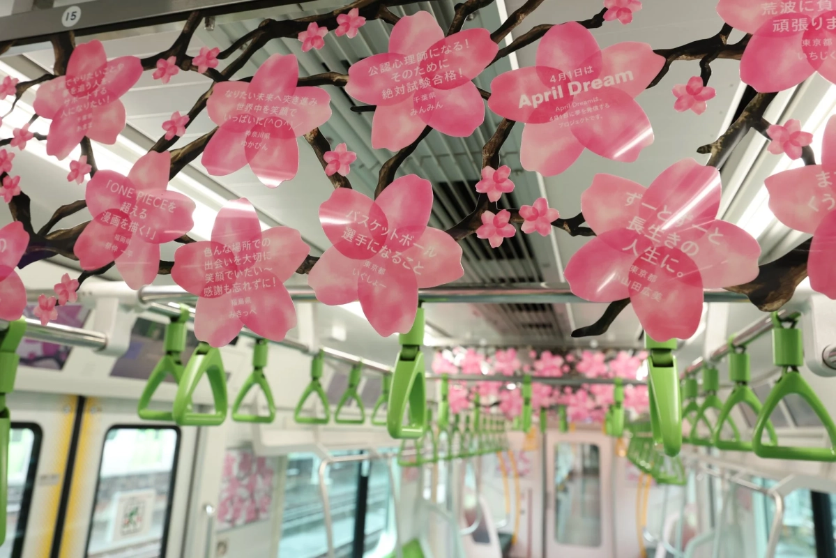 電車内に桜の形をしたピンク色の装飾が吊り下げられており、それぞれに「April Dream」プロジェクトの一環として人々の夢や願いが書かれている。緑色のつり革が並び、春らしい華やかな雰囲気の車内風景。