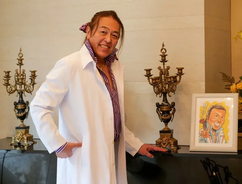 白いコートとストライプシャツを着た男性が、笑顔でカメラにポーズをとっている