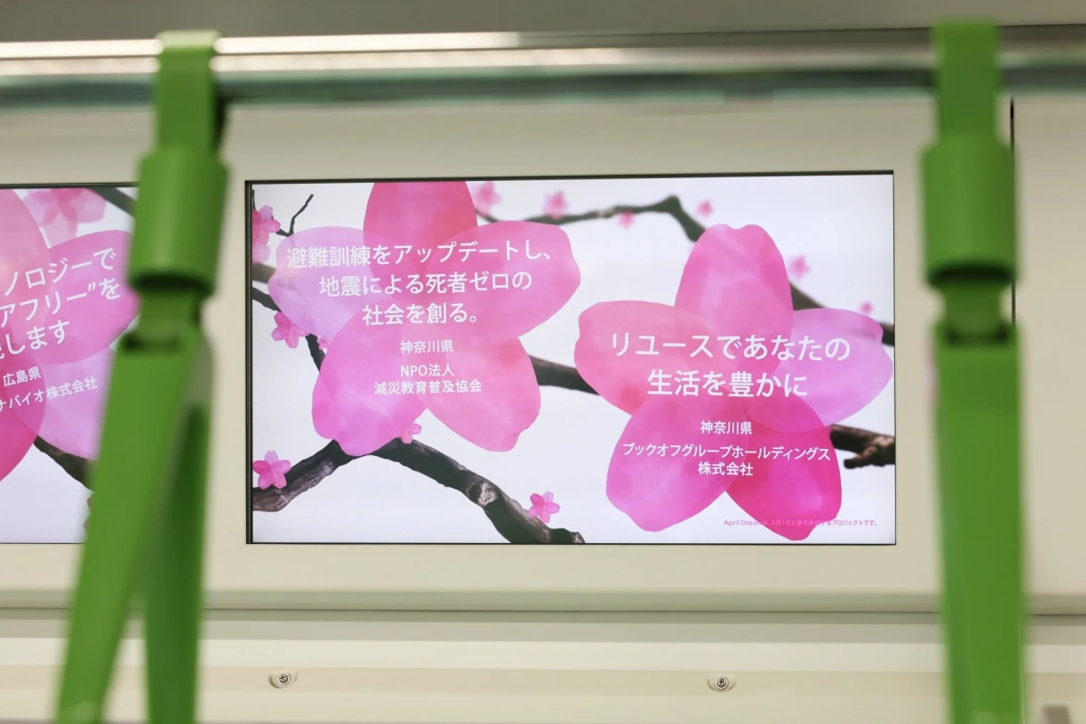 電車内のデジタルサイネージに表示された広告で、桜の花びらを背景に、地震による死者ゼロを目指す減災教育普及協会と、リユースで生活を豊かにするブックオフグループホールディングスの取り組みが紹介されています。April Dreamプロジェクトの一環です。