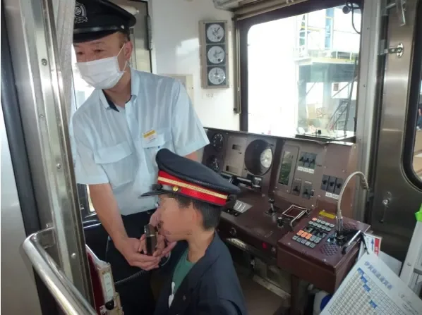 電車の運転席で、マスク姿の運転士が子供に運転操作を教えている様子