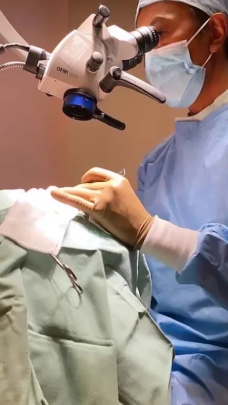 手術着を着た外科医が、手術用顕微鏡を覗き込みながら、手袋をはめた手で精密な医療処置を行っている様子