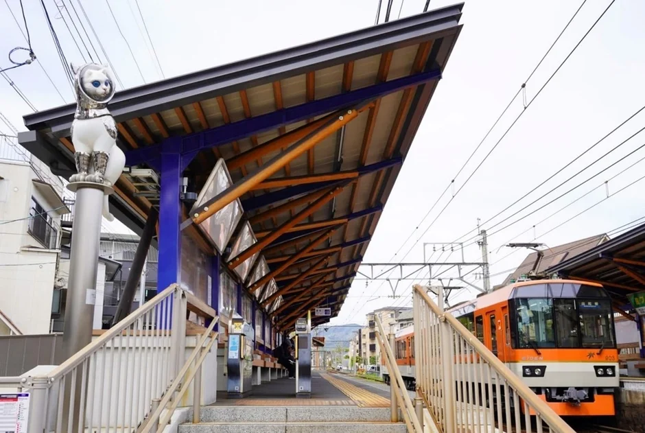 宇宙服姿の猫の像が立つユニークな鉄道駅のプラットフォームに、オレンジ色の電車が停車しています。モダンなデザインの駅舎が特徴的です。