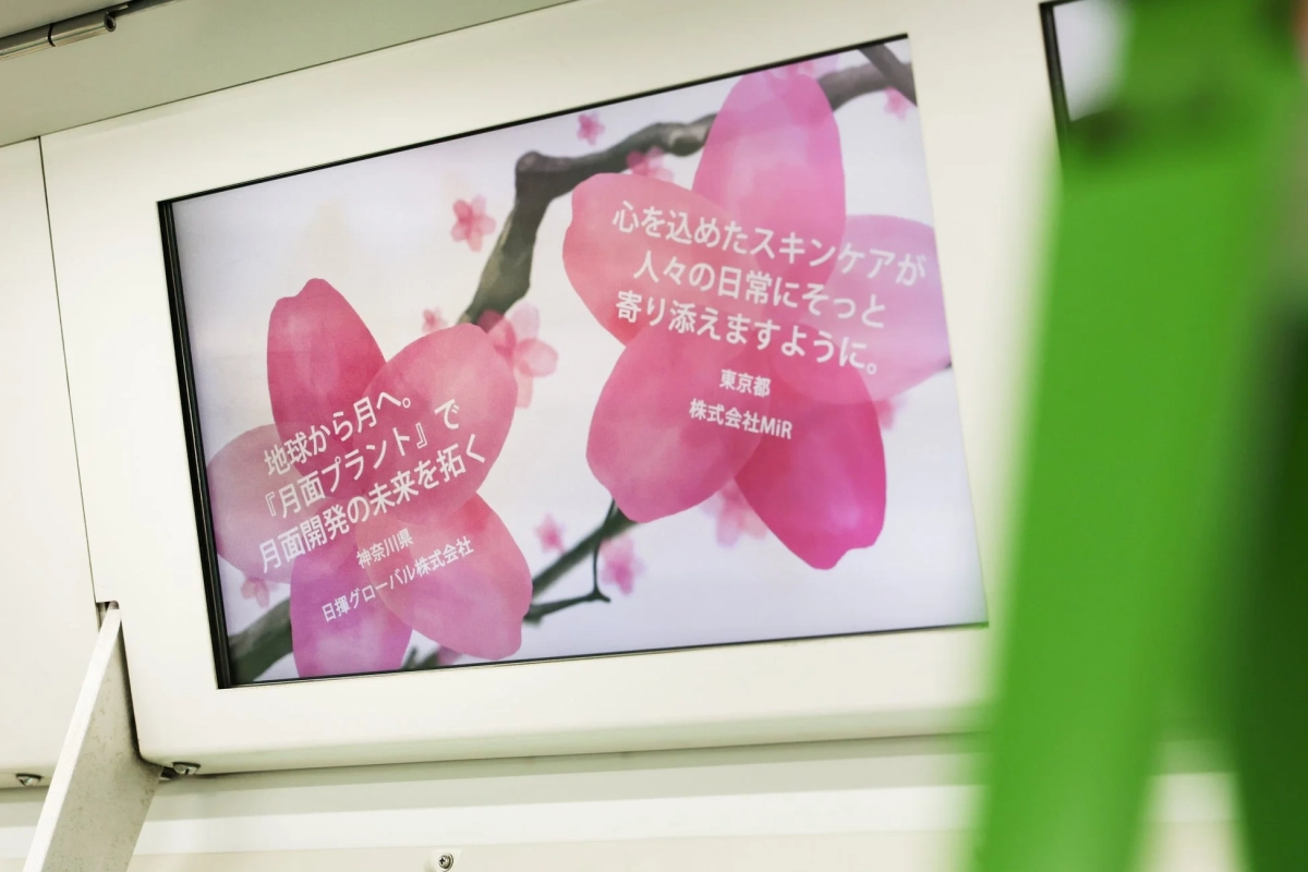 電車内のデジタル広告画面に、桜の花を背景にした2つの企業広告が表示されています。一つは「心を込めたスキンケア」を、もう一つは「月面プラントで月面開発の未来を拓く」という、異なる分野のメッセージが並列に示されています。