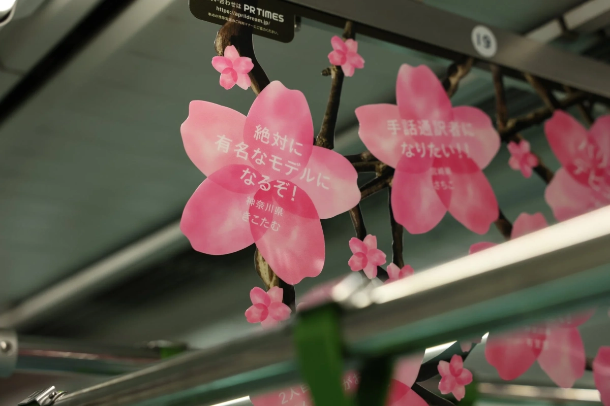 電車内らしき空間に、桜の花びらを模したピンク色の広告が吊るされています。広告には「有名なモデルになるぞ!」「手話通訳者になりたい!」といった、人々の夢や目標が書かれており、春の訪れと共に希望を感じさせるデザインです。