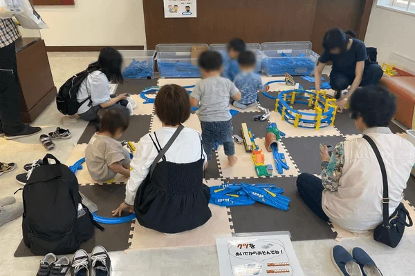 子供たちが大人と一緒に室内でプラレールの電車のおもちゃで遊んでいる様子
