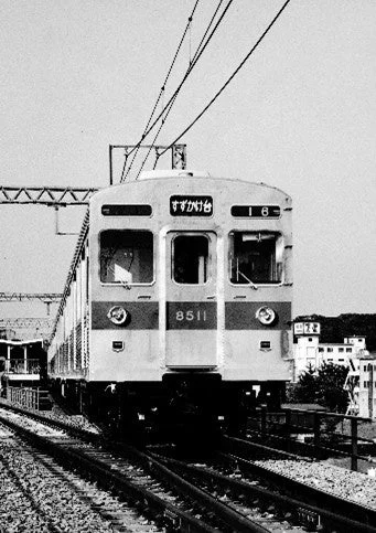 鉄道の線路上を走る電車の白黒写真。電車の前面には行き先表示「すずかけ台」と車両番号「8511」が読み取れる。架線と線路が印象的な、ノスタルジックな鉄道の情景を捉えている。