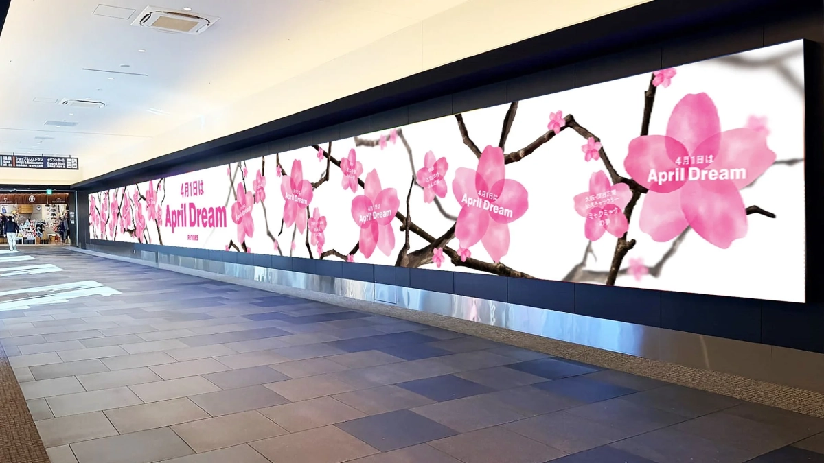 商業施設の通路に設置された大型デジタルサイネージに、ピンク色の桜と「4月1日は April Dream」の文字が表示されています。PR TIMESによる夢をテーマにしたキャンペーンの告知で、春らしい華やかな雰囲気です。