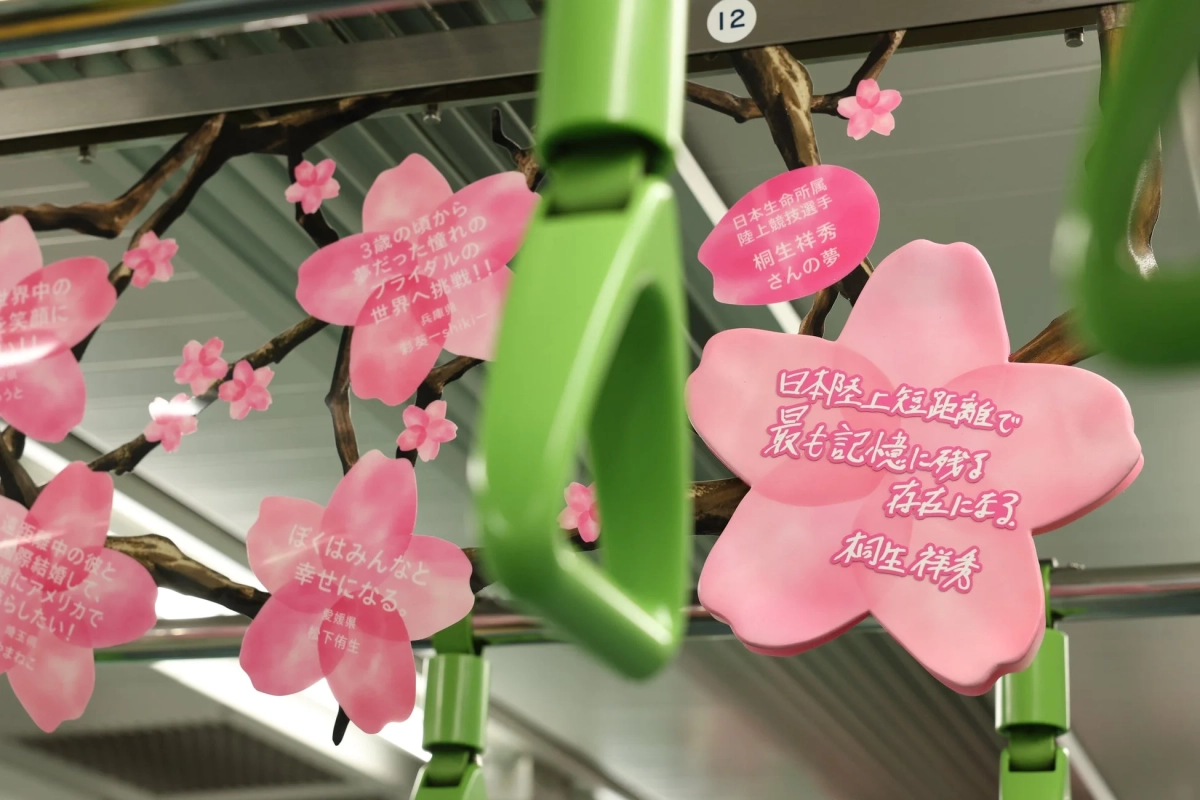 電車の車内が多数のピンクの桜の飾りで装飾されており、緑色の吊り革が手前に見える。桜の花びらには、陸上競技選手の夢や、ブライダル、国際結婚、幸せに関する様々な個人のメッセージが書かれている。