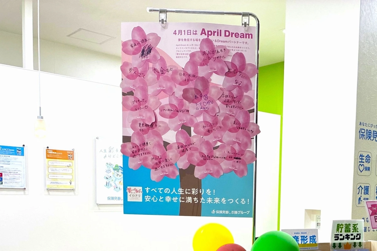 「April Dream」と題された桜の木を模した掲示物が写っています。ピンク色の桜の花びらには、参加者それぞれの夢や願い事が書かれており、健康や大金持ち、旅行、特定の職業になることなど、様々な目標が表現されています。このイベントは、夢を通じて未来を創造することを目的としており、「保険見直し本舗グループ」が関わっているようです。全体的に明るく前向きな雰囲気が伝わります。