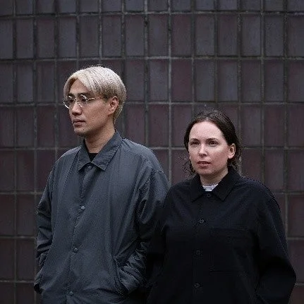 金髪の男性と黒髪の女性が並んで立つポートレート写真です。男性はメガネをかけ、ダークグレーのジャケットを着用しています。女性は黒いジャケットを着ており、二人とも落ち着いた表情をしています。背景は暗い壁で、都会的な雰囲気を醸し出しています。