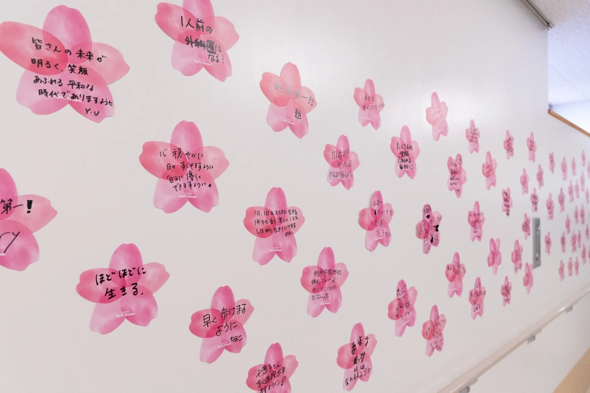白い壁に桜の形をしたピンクの付箋が多数貼られており、それぞれに手書きのメッセージが書かれています。未来への希望、平和、健康、夢、応援など、様々な願いが込められた温かいメッセージで壁が彩られています。