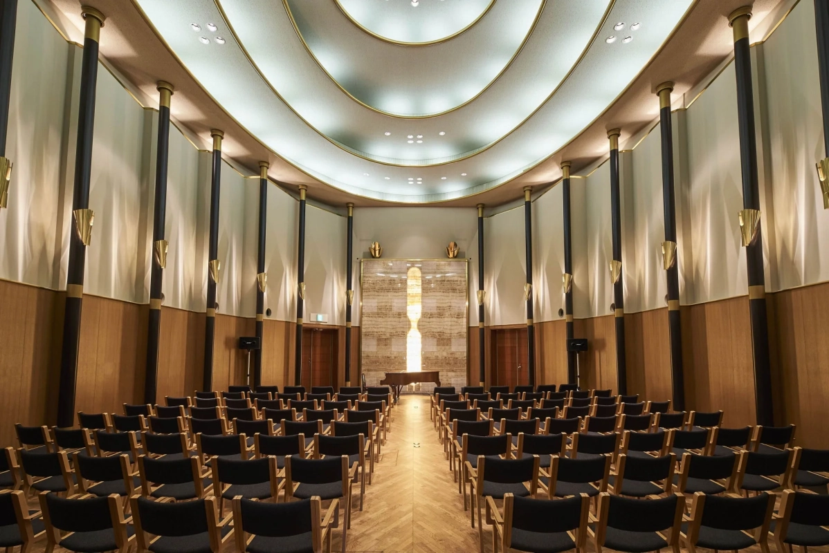 モダンなデザインの講堂またはイベントホールで、整然と並べられた椅子とステージに置かれたピアノが見える