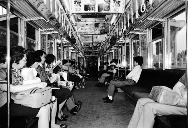 昭和時代の日本の電車内を写した白黒写真。多くの乗客が座席に座り、読書をしたり、静かに過ごしたりしている様子がうかがえ、当時の日常的な移動風景を伝えている。