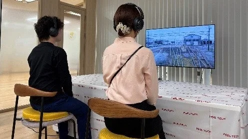 ヘッドホンを装着した男女2人が、鉄道風景が映し出された大型ディスプレイを熱心に見ている様子