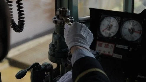 白い手袋をはめた運転士が、計器類と操作レバーのある運転席で機器を操作している様子