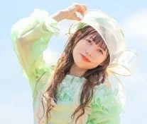 明るい青空を背景に、緑色の服と透明な帽子を身につけた若い女性が笑顔でこちらを見ているポートレート写真