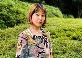 緑豊かな自然を背景に、ショートヘアの女性がカメラを見つめて立っています。柄物のトップスを着用しており、穏やかな表情です。