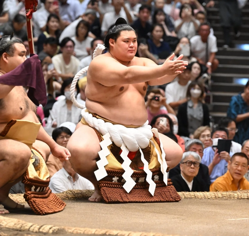 大相撲の土俵で、横綱が蹲踞の姿勢で手を叩く準備をしている様子。豪華な化粧まわしを身につけ、多くの観客が見守る中、伝統的な儀式が執り行われています。