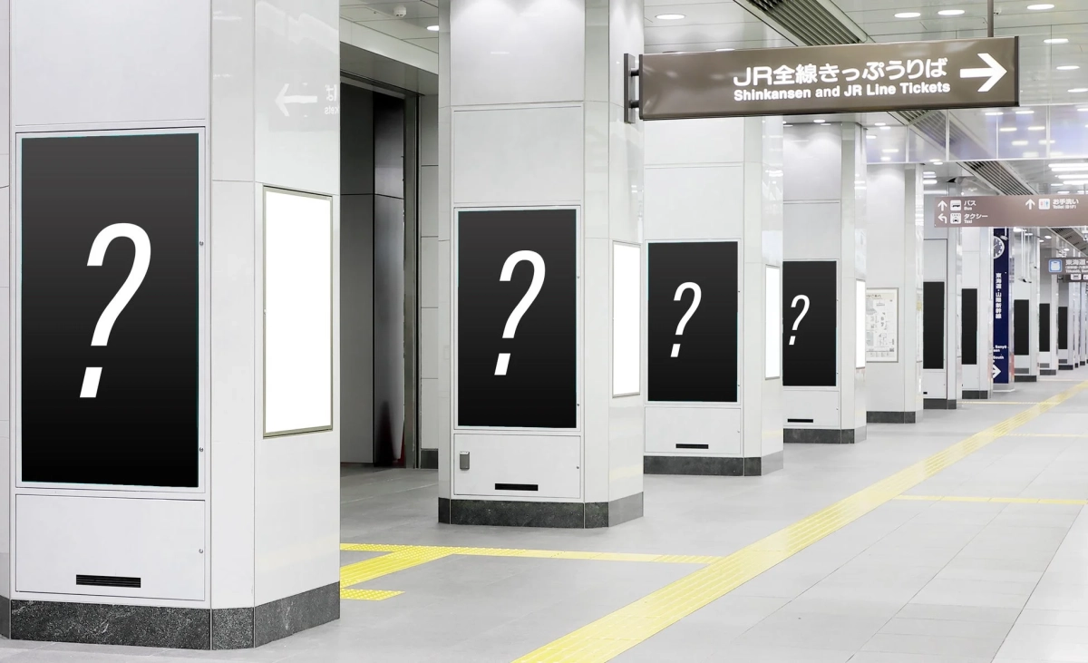 日本の駅構内または地下通路のような場所で、白い柱が規則的に並んでいます。柱には広告ディスプレイが設置されており、多くに白い疑問符が表示されています。JR全線きっぷうりばへの案内標識も見え、清潔でモダンな印象の空間です。