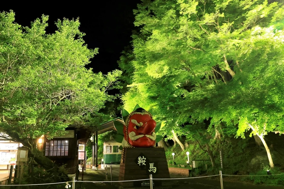 夜の風景で、巨大な赤い天狗の像が緑にライトアップされた木々の間に立っています