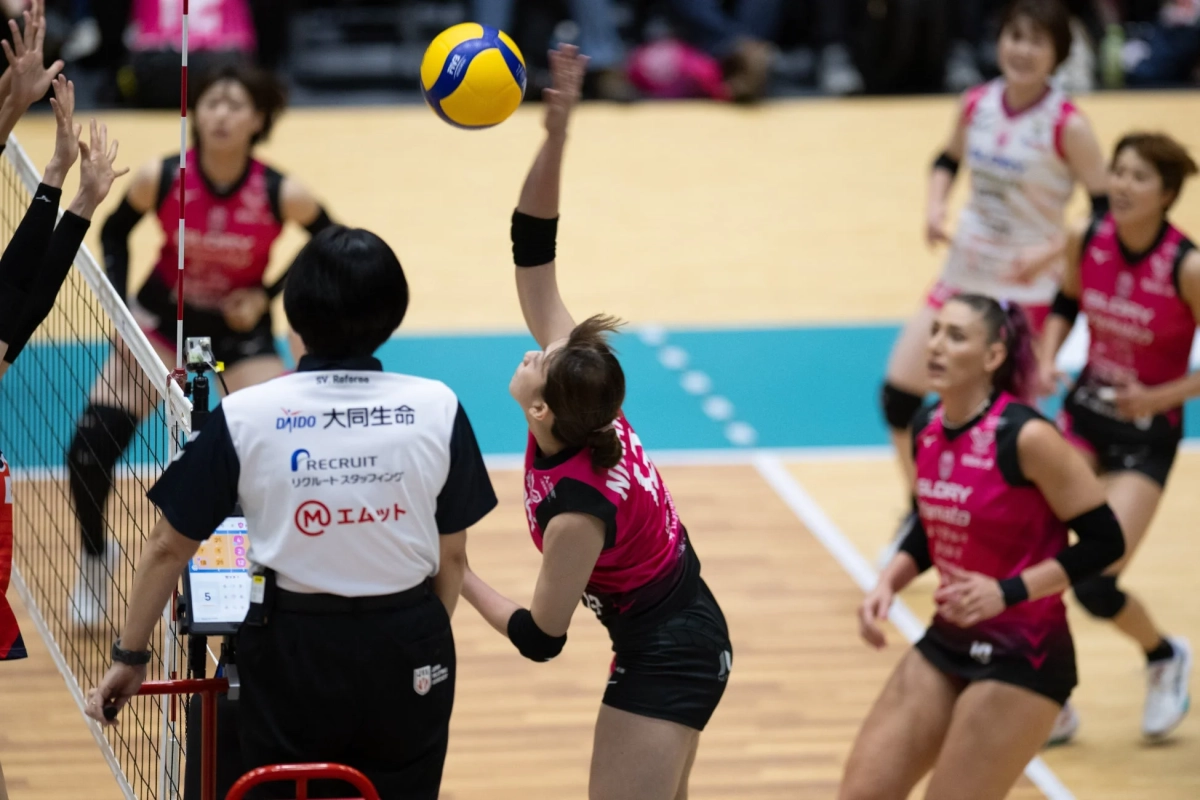 バレーボールの試合で、ピンクのユニフォームを着た選手がボールを打ち上げる瞬間の躍動感ある一枚。ネット際では審判がスコアボードを確認しており、他の選手たちも真剣な表情でプレイを見守る様子が写し出されています。