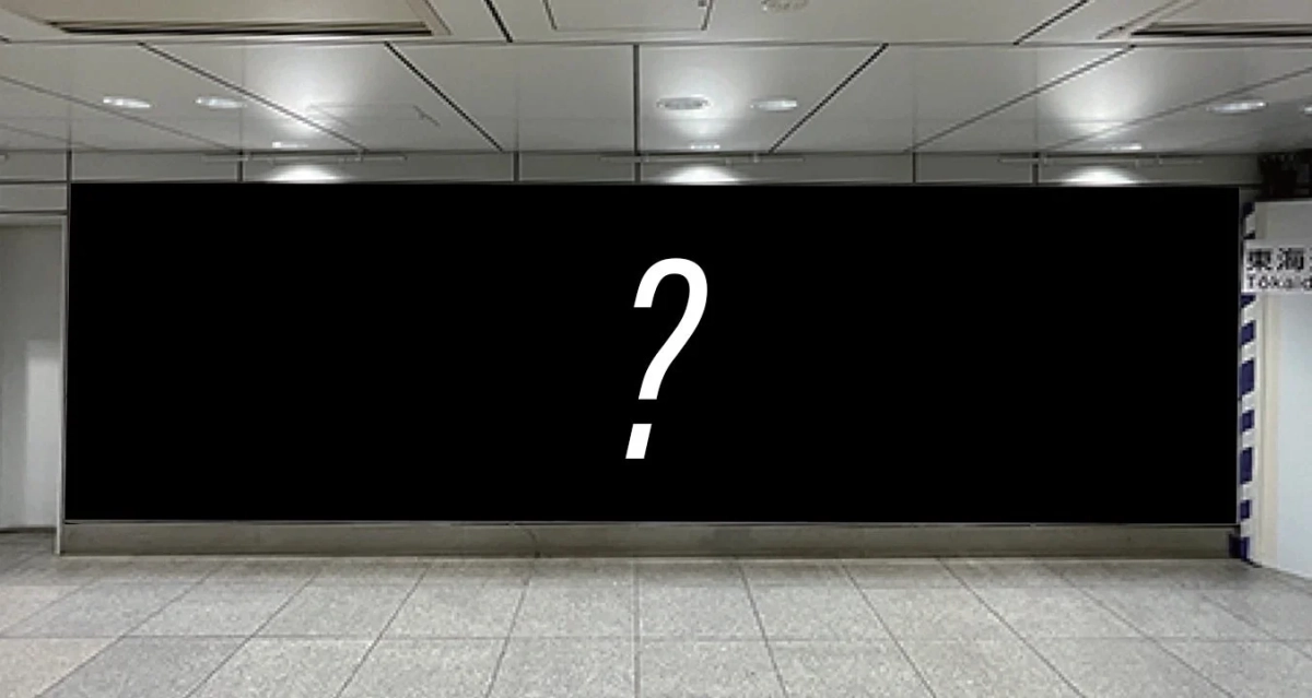 駅構内のような屋内の通路に設置された大きな黒い広告板です。中央には白い大きな疑問符が表示されており、右端には「東海 Tōkaido」と書かれた案内板の一部が見えます。何か新しい情報が始まる前の期待感や未定の状態を示唆しています。