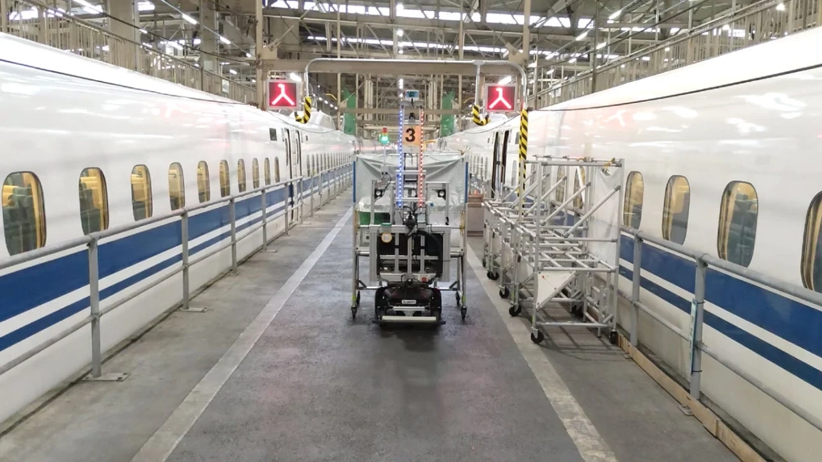 新幹線車両基地の内部で、2台の新幹線が並んで停車しており、その間には自動点検ロボットのような機器が設置されています。