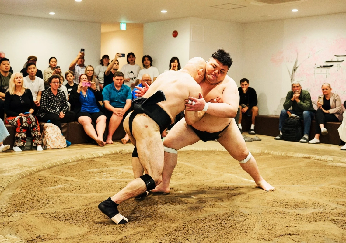 土俵の上で二人の力士が相撲を取っており、多くの観客がその様子を見守っています。日本の伝統的なスポーツイベントの瞬間を捉えたものです。