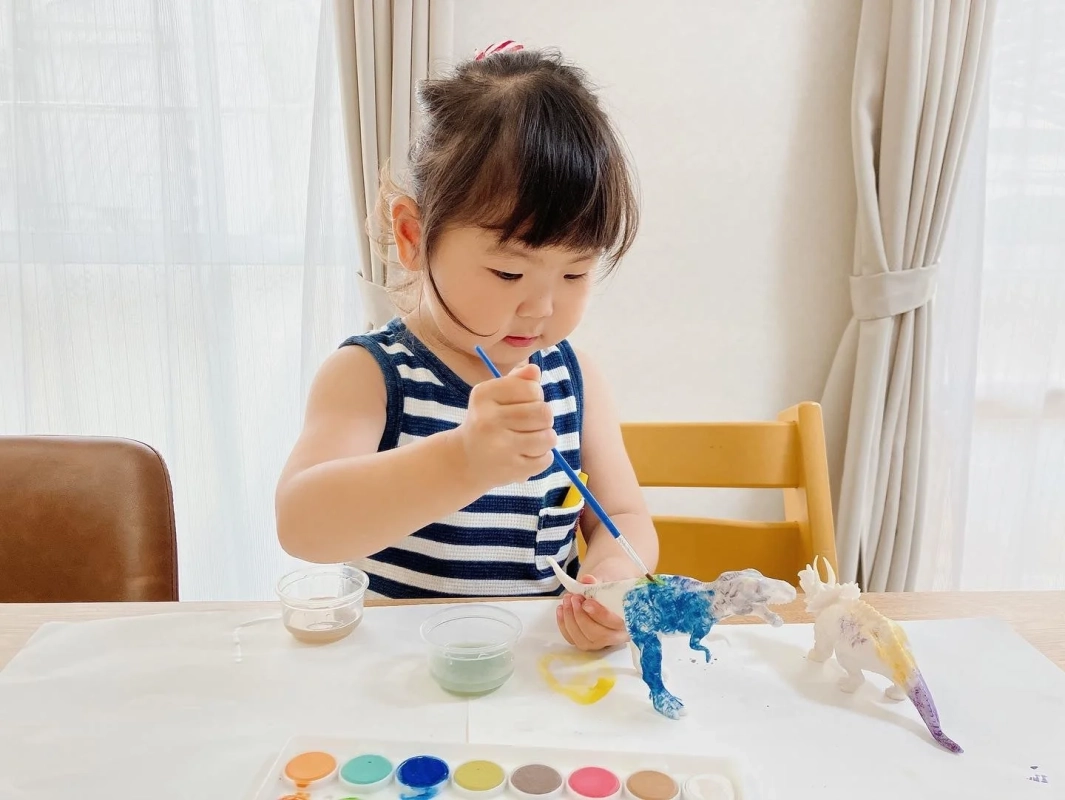 小さな女の子がテーブルで恐竜のフィギュアに水彩絵の具を使って色を塗っている